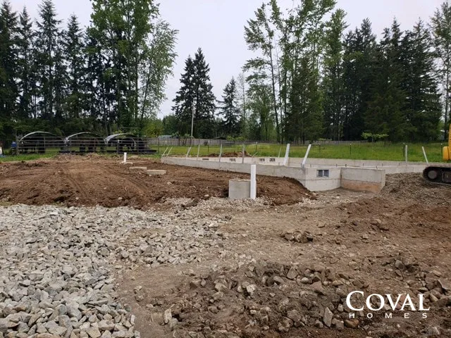 Construction site with gravel piles, concrete footings, and trees in the background