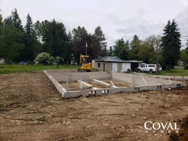 Concrete foundation under construction beside a wooded lot, with an excavator and truck nearby