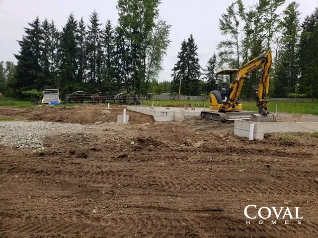 Construction site with yellow excavator leveling dirt near concrete foundations and trees in the background