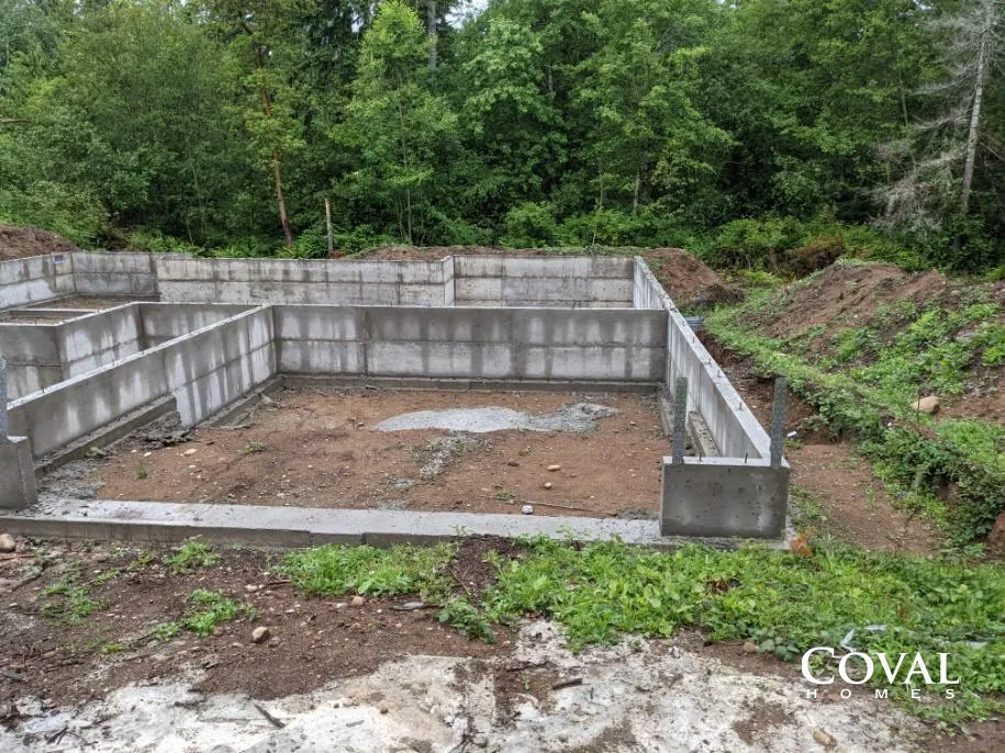 Concrete foundation walls on a muddy site beside dense green trees