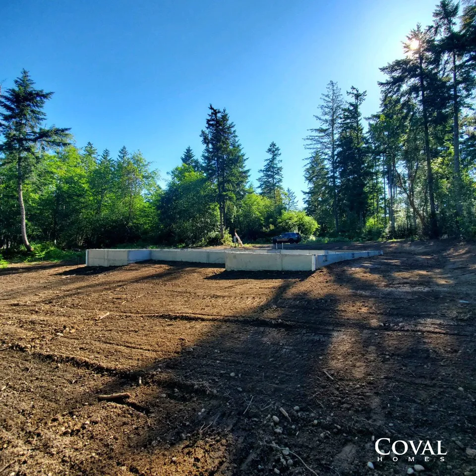 Cleared dirt lot beside a low concrete wall, surrounded by tall pine trees under a bright blue sky.