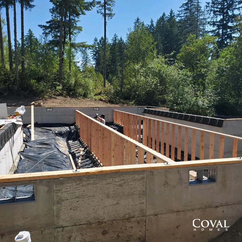 Construction site with concrete foundation walls and wooden framing under a sunny forested sky