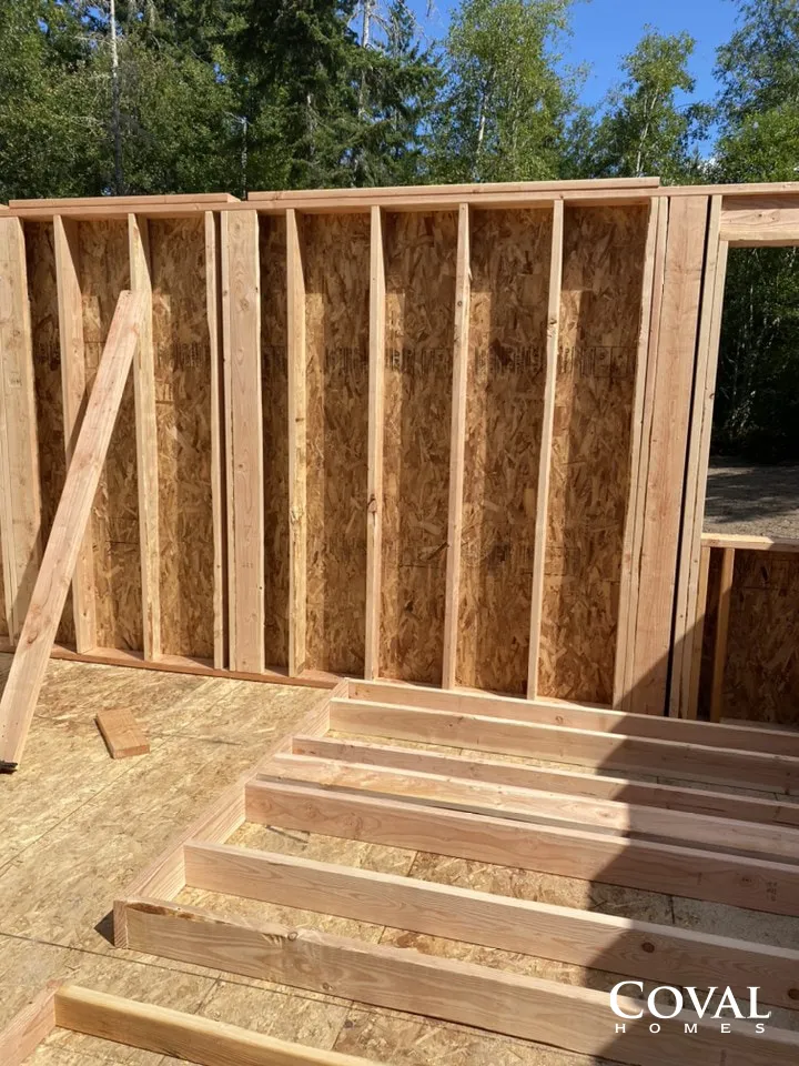 Framed wooden wall under construction with studs and roofline, lumber stacked in the foreground outdoors