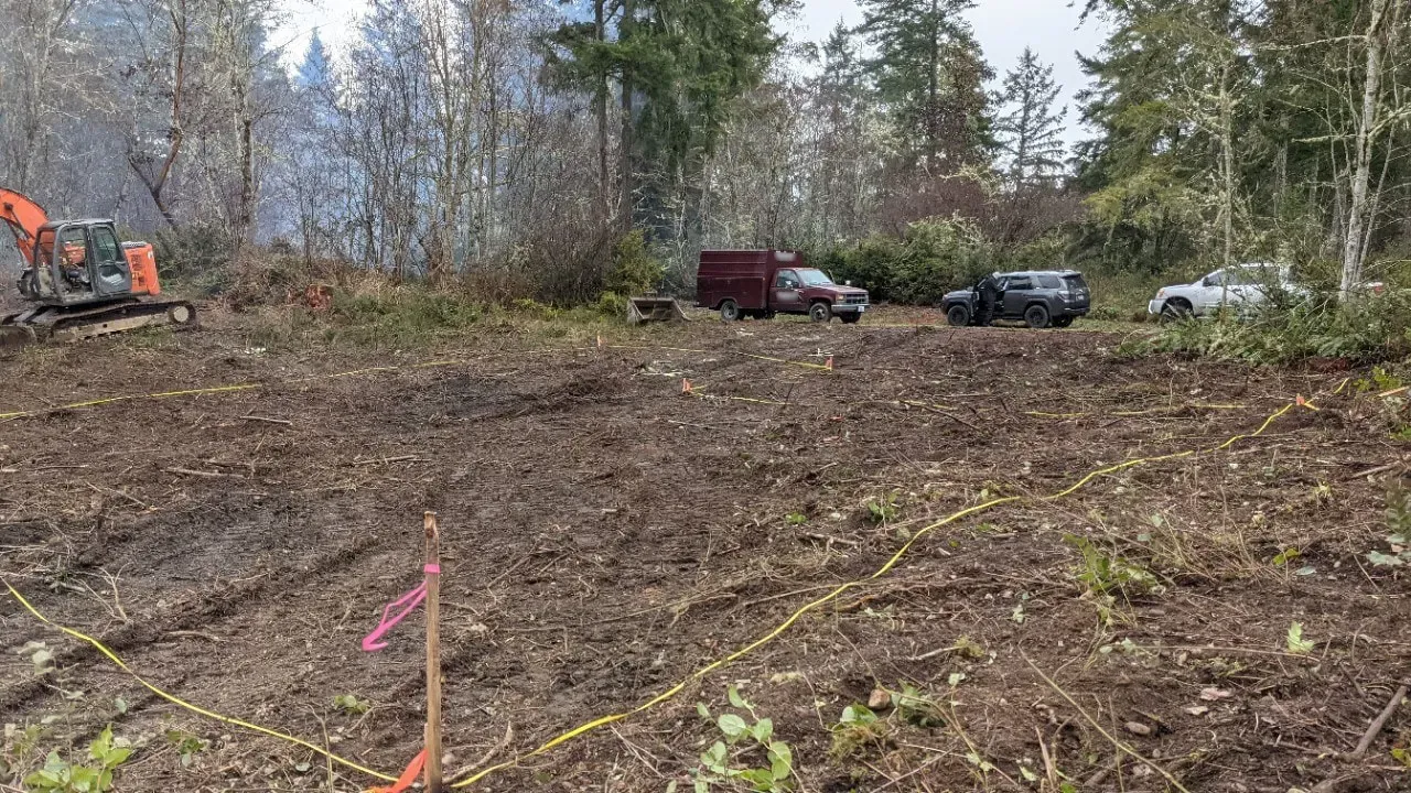 Cleared muddy lot with excavator, parked vehicles, and trees in the background