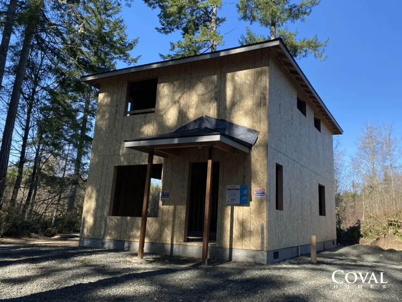 Two-story wooden cabin under construction in a wooded area with a gravel driveway.