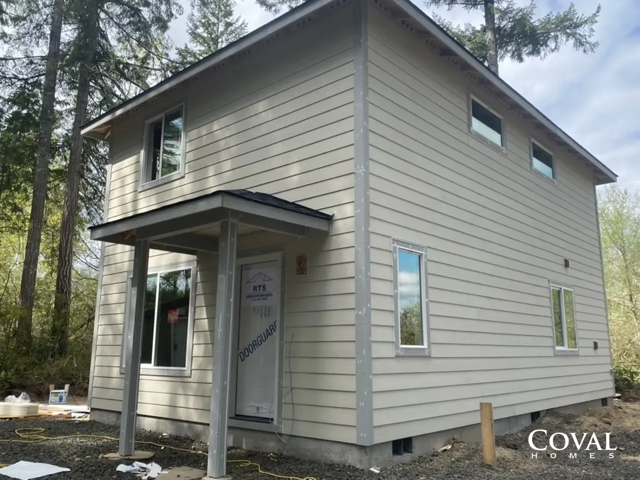 Small beige two-story house under construction with a front porch, surrounded by trees.