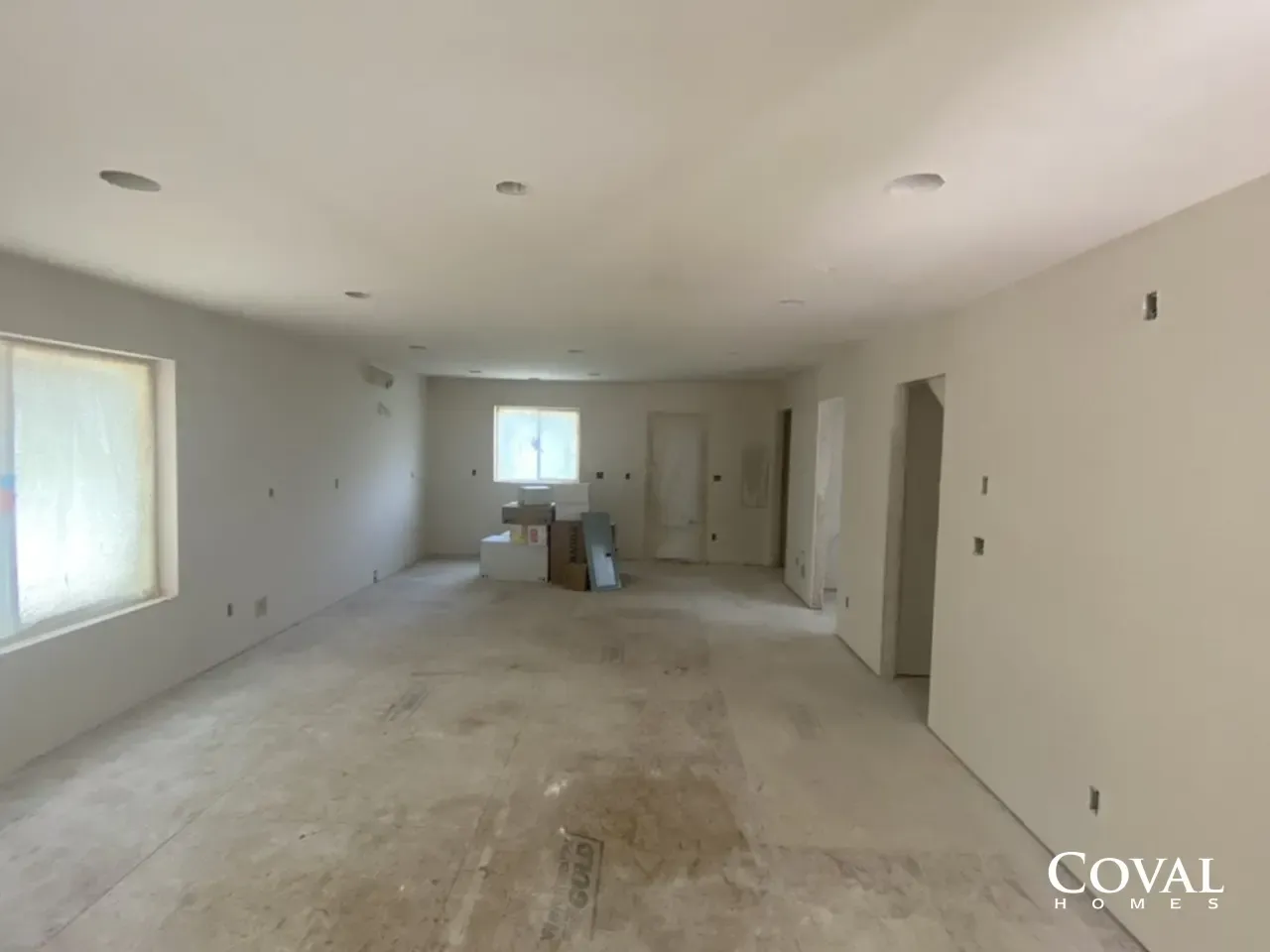 Empty unfinished room with white walls, recessed lights, and windows, covered in dust and construction debris.