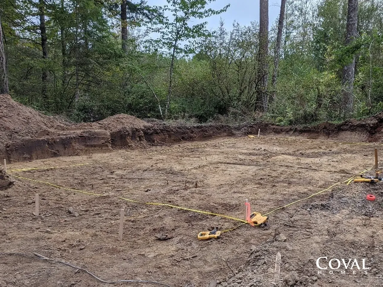 Cleared dirt lot with wooded edge and survey markers under construction on a cloudy day