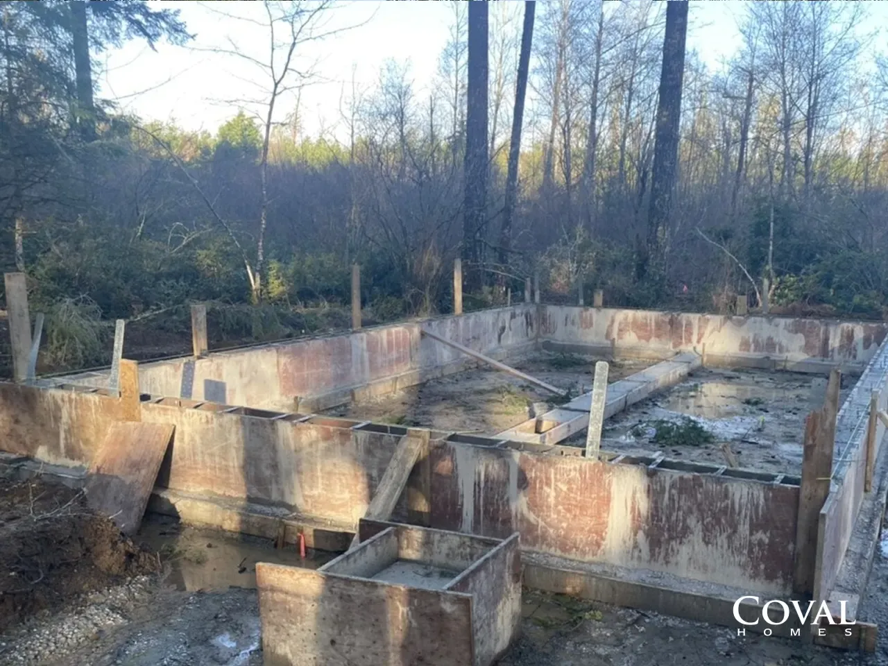 Abandoned concrete foundation in a wooded area, with low walls and trees in the background.