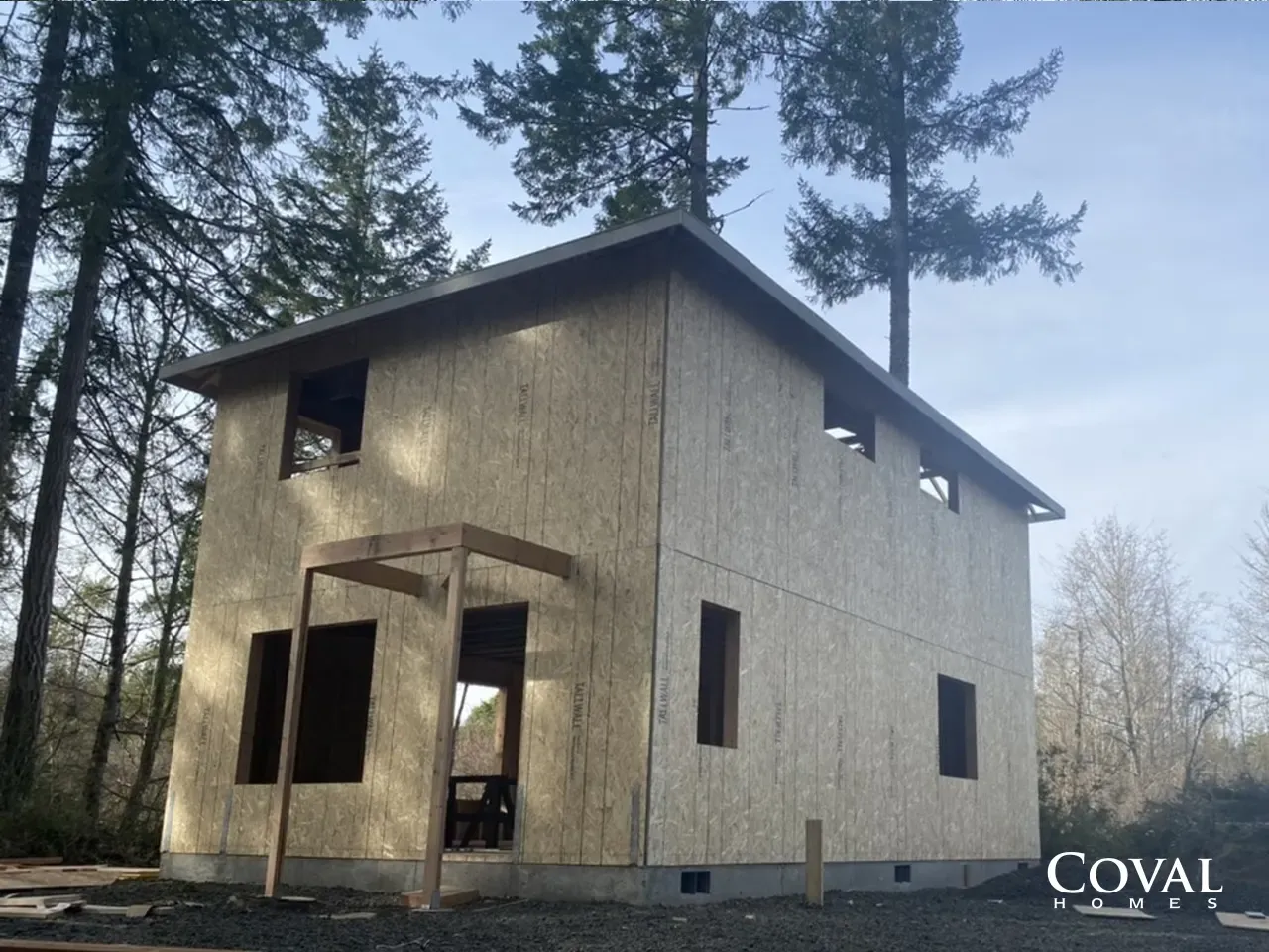 Small unfinished two-story beige house in the woods, with a covered porch and tall trees around it.