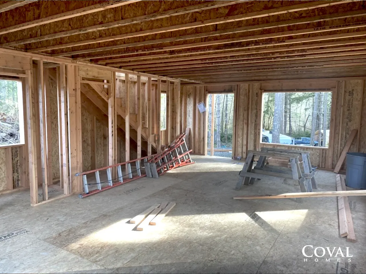 Unfinished wooden house interior with exposed framing, ladders, and sunlight through open windows.
