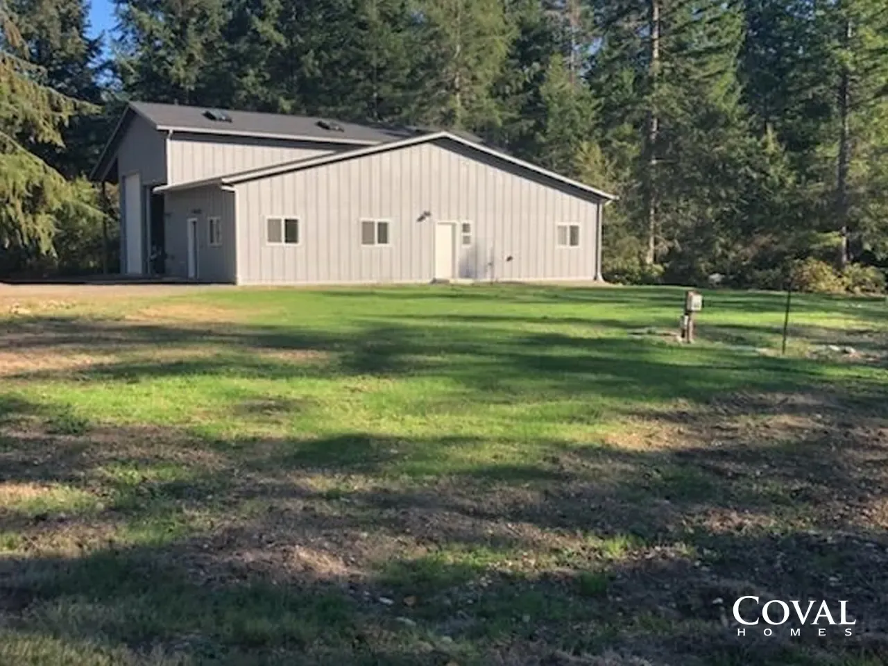 Beige metal barn on a grassy yard with trees in the background and a small dog near the right side
