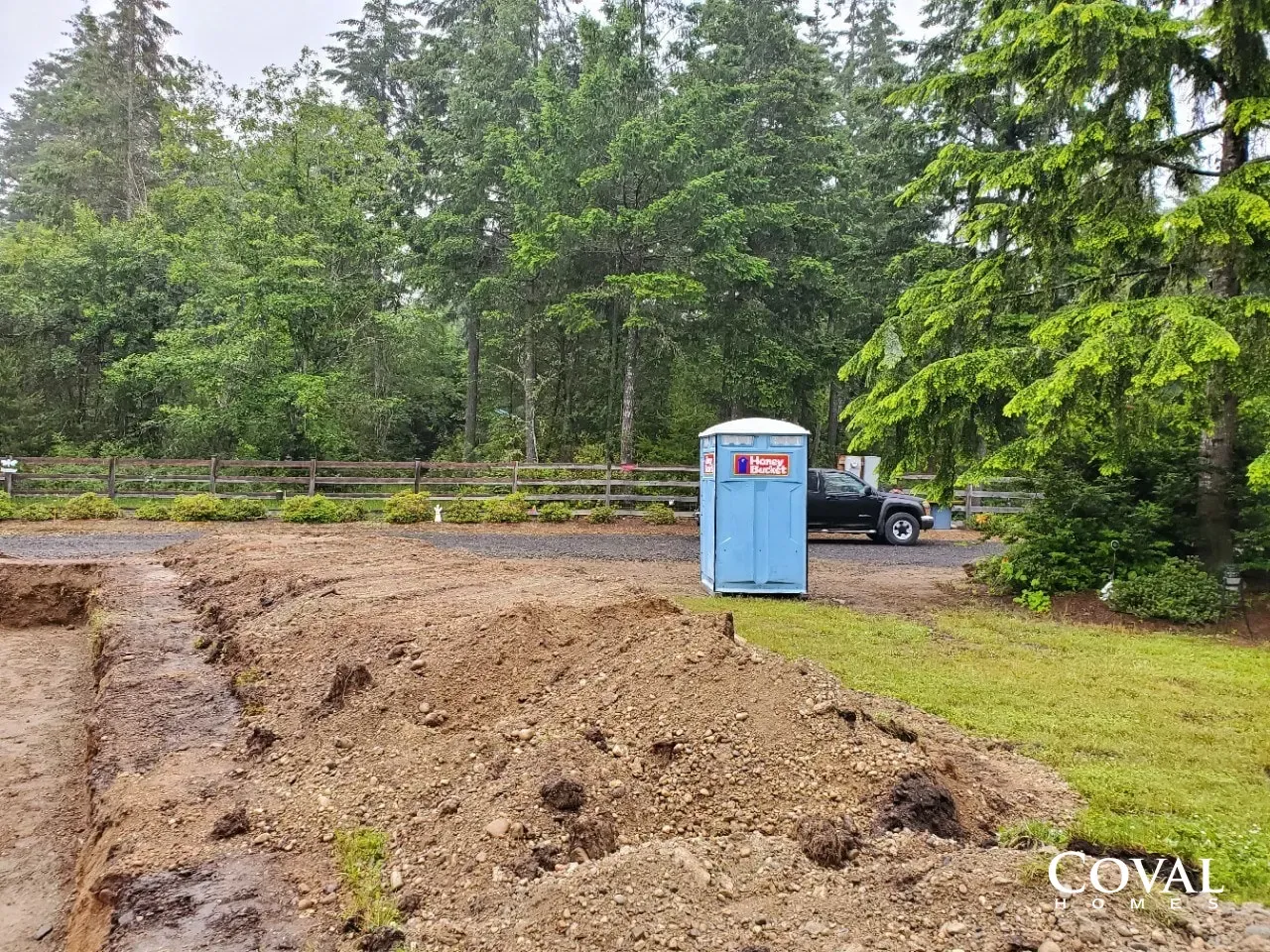 Blue portable toilet beside muddy construction site, with a pickup truck and trees in the background