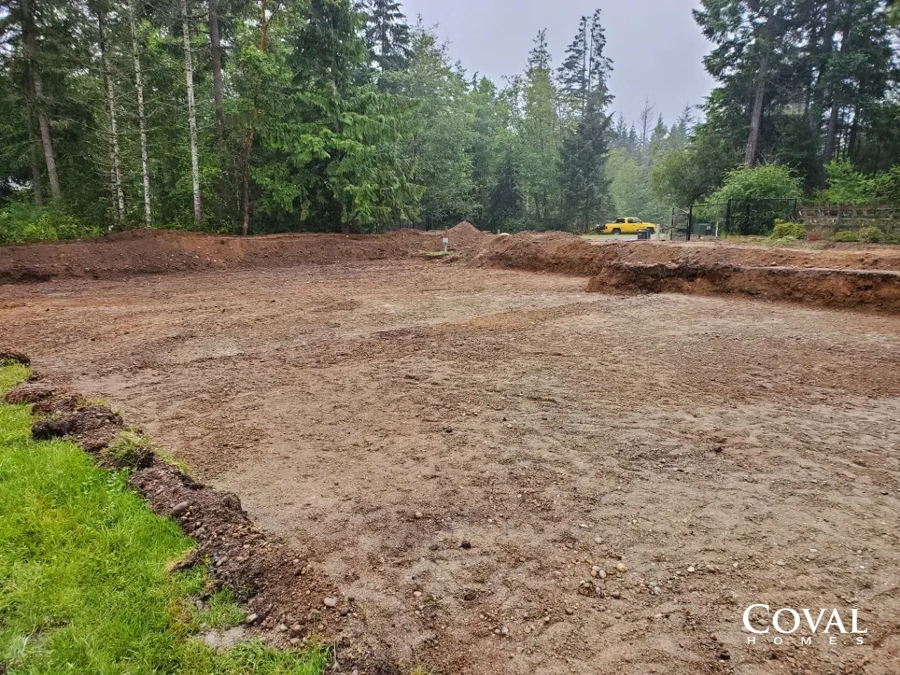 Cleared dirt lot bordered by trees, with a small excavator in the distance