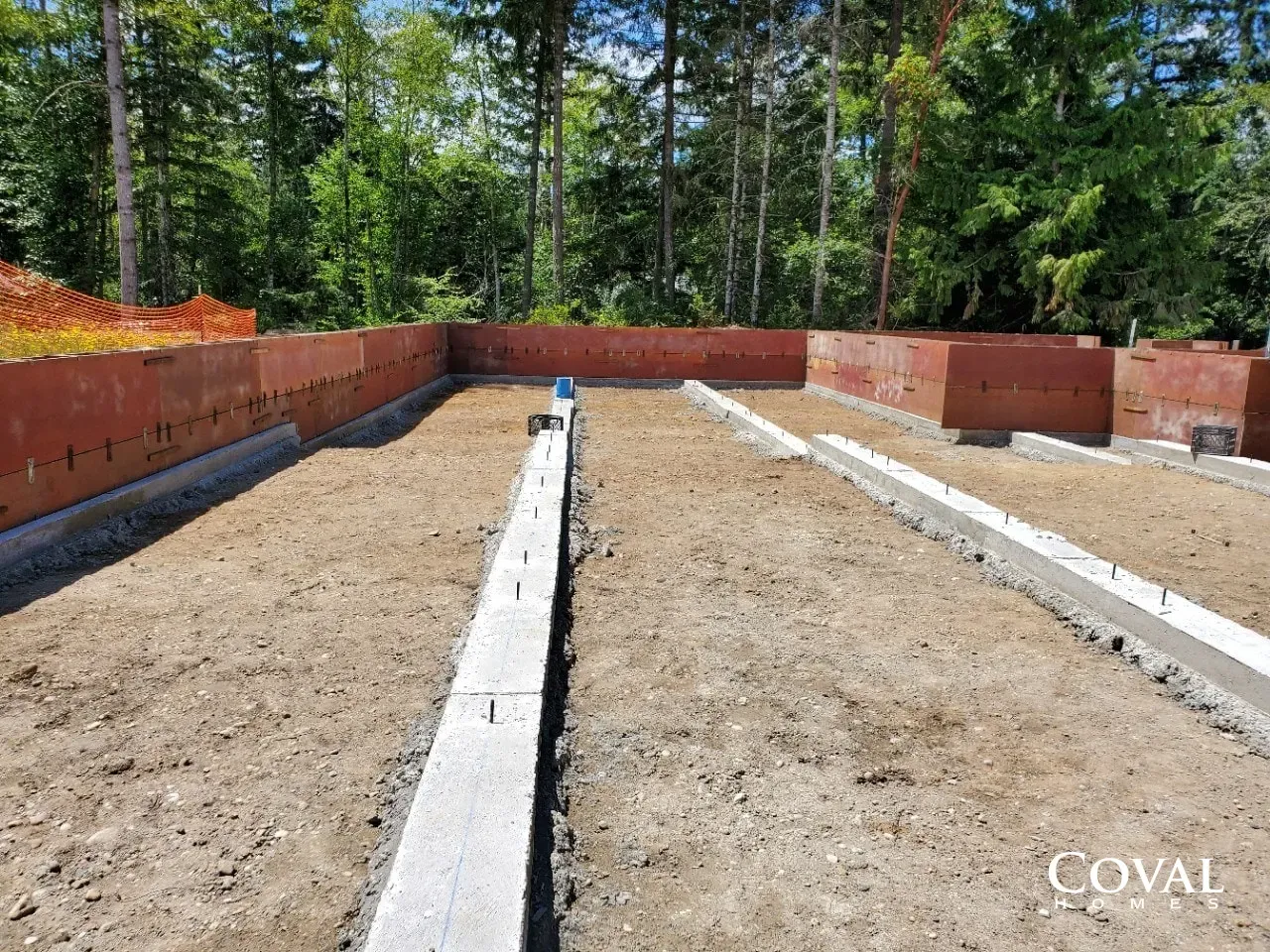 Concrete foundation footings in a cleared lot, with red retaining walls and trees in the background.