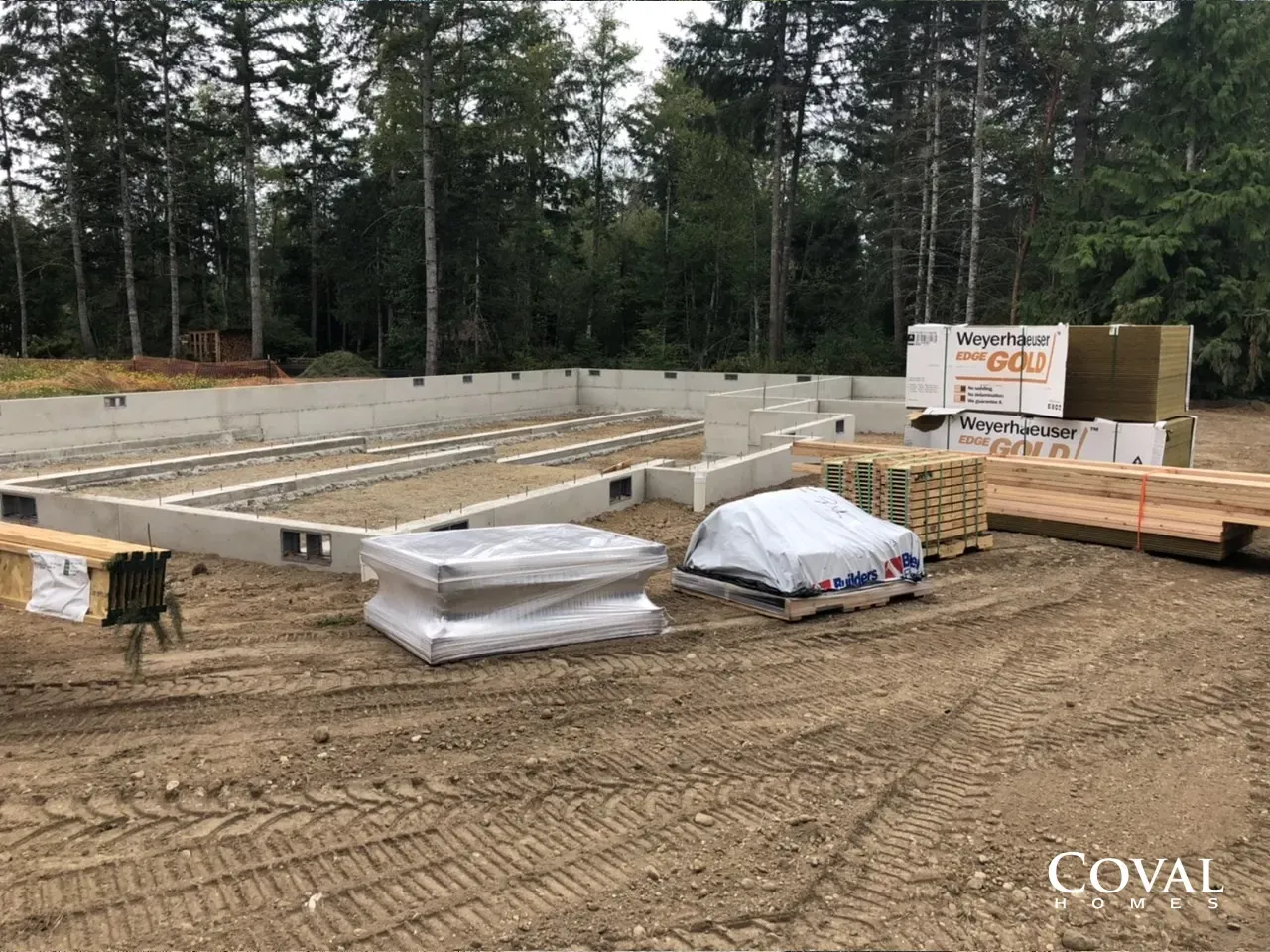 Foundation under construction with concrete footings, lumber stacks, and forested background