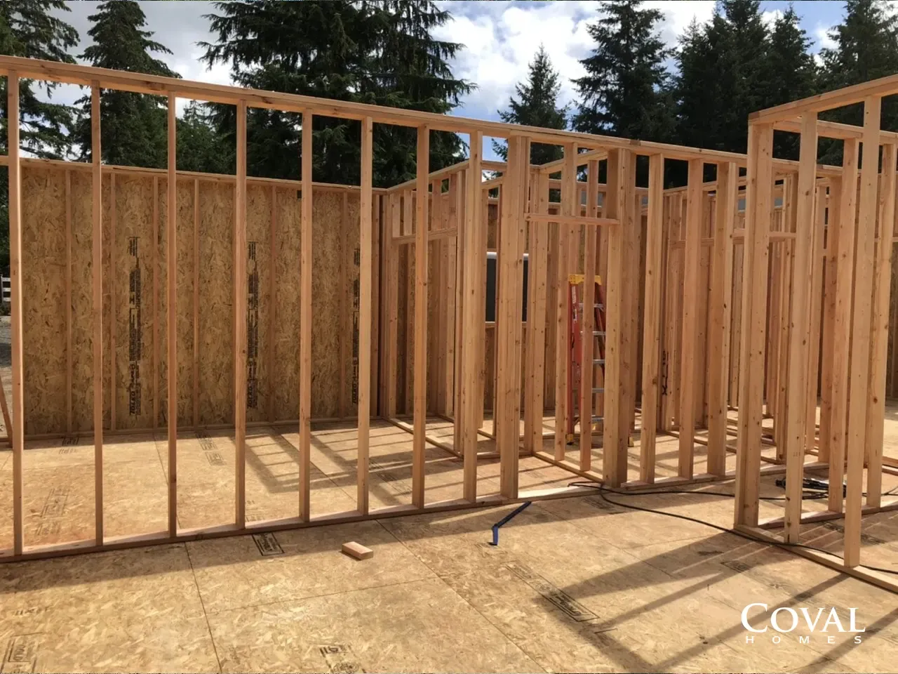 Wooden house framing under construction on a sunny day, with exposed studs and foundation slab.