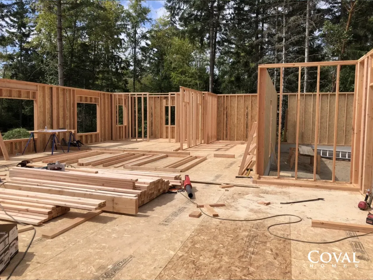 Wood-framed house under construction with lumber stacks and forest background