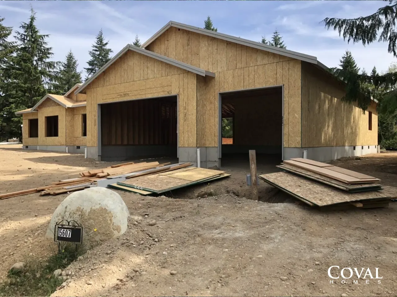 Wood-framed barn under construction with open bays, plywood ramps, and dirt lot surrounded by trees