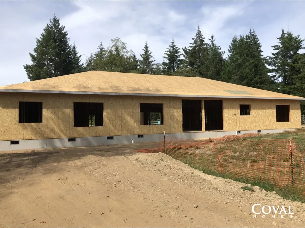 Unfinished single-story house with tan siding and dark window openings, surrounded by dirt and trees.