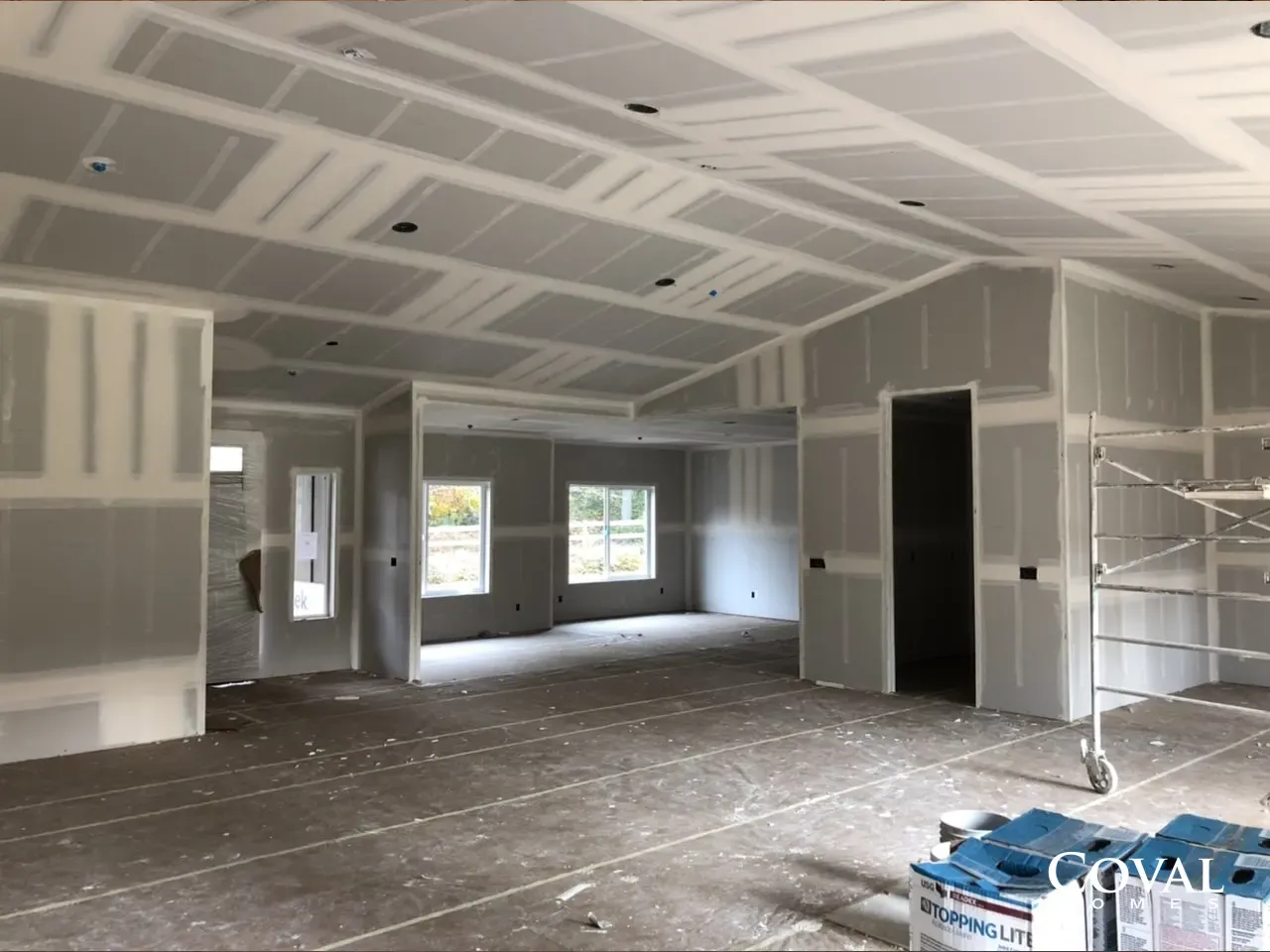Unfinished drywall interior with exposed ceiling, windows, and construction materials on the floor
