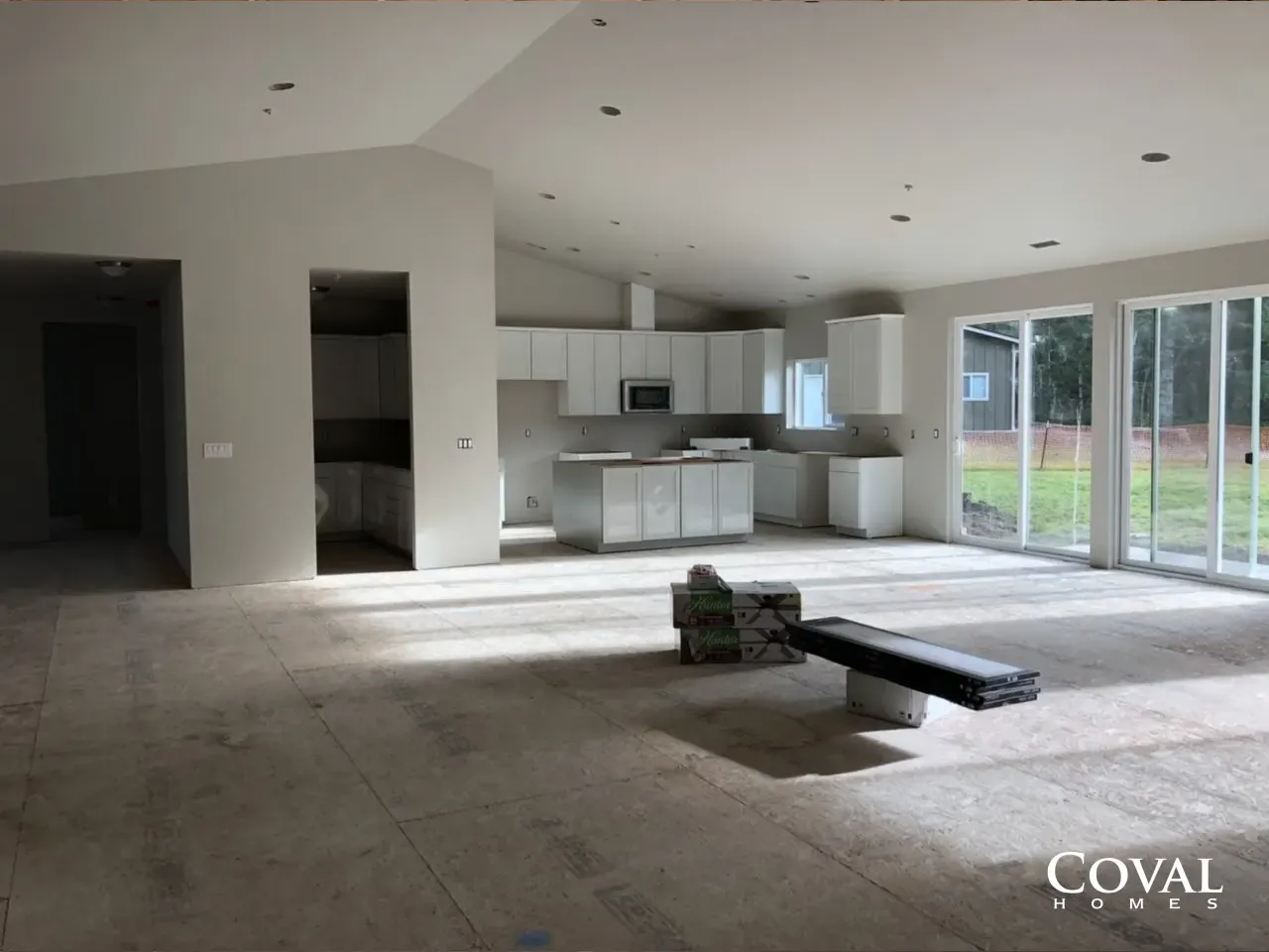 Empty modern living room with unfinished floor, white kitchen, and large glass doors to a yard