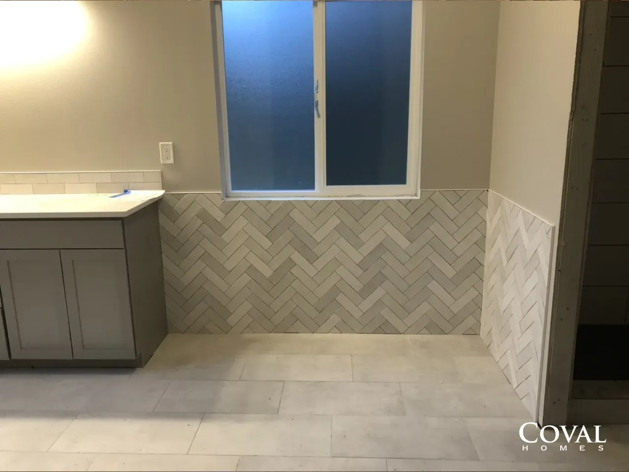Empty bathroom with gray herringbone tile, white vanity, and a window above the tub area.