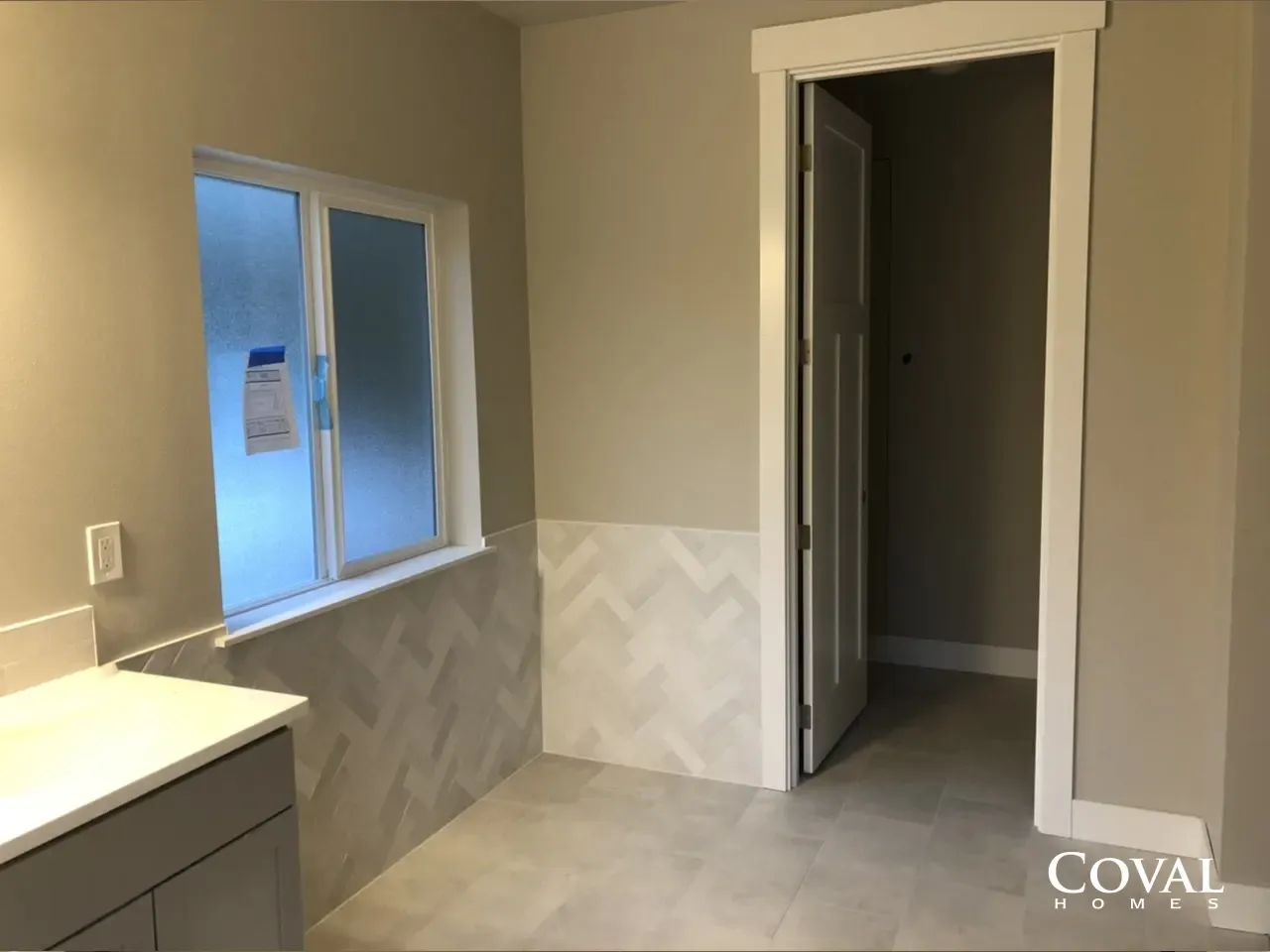 Empty beige bathroom with vanity, window, tiled wainscoting, and open doorway to a hallway