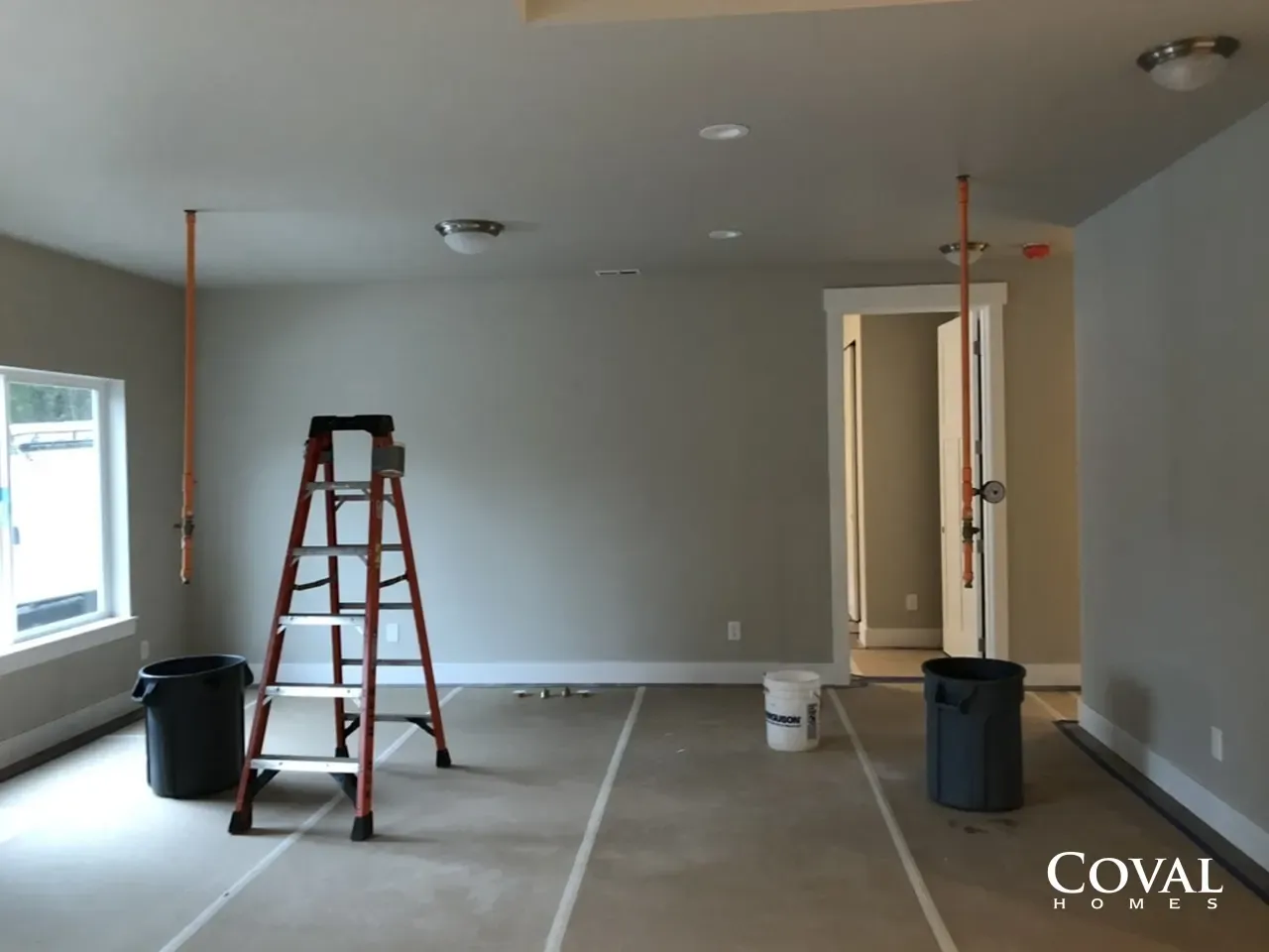 Empty room under renovation with ladder, paint bucket, and trash cans on a concrete floor.