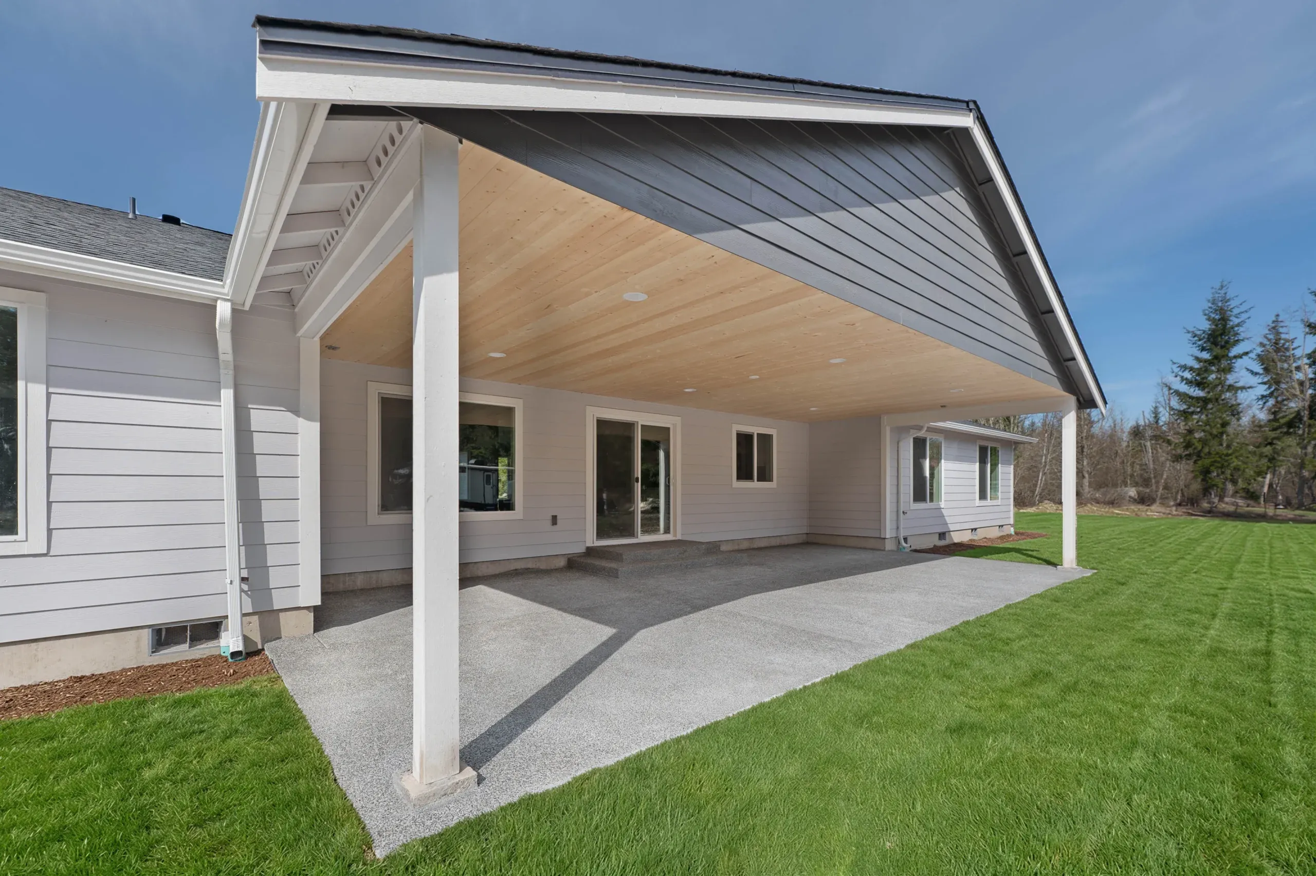 Covered patio attached to a modern white house, with gravel flooring and a green lawn under a blue sky.
