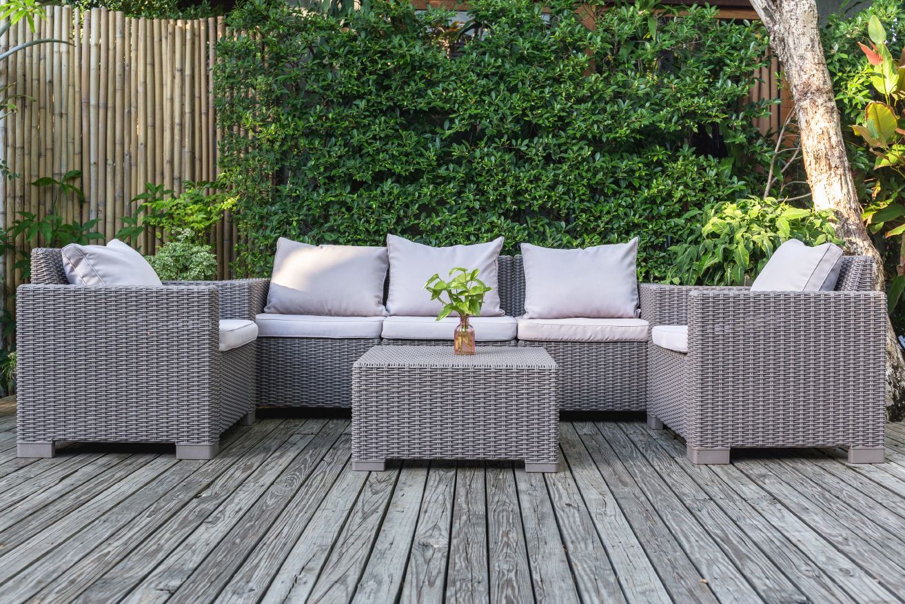 Modern gray wicker patio seating with cushions and a small table on a wooden deck, backed by greenery