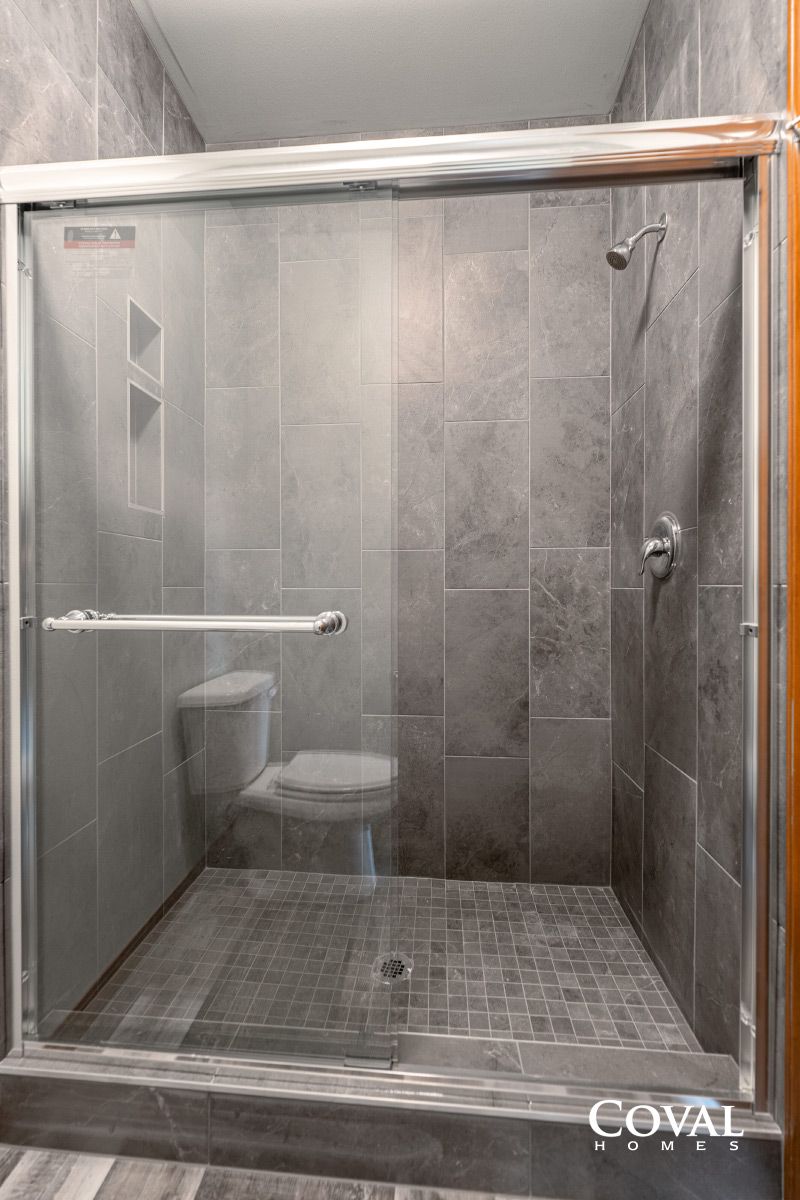 Accessible walk-in shower with bench, toilet, grab bar, and glass door in a gray-tiled bathroom