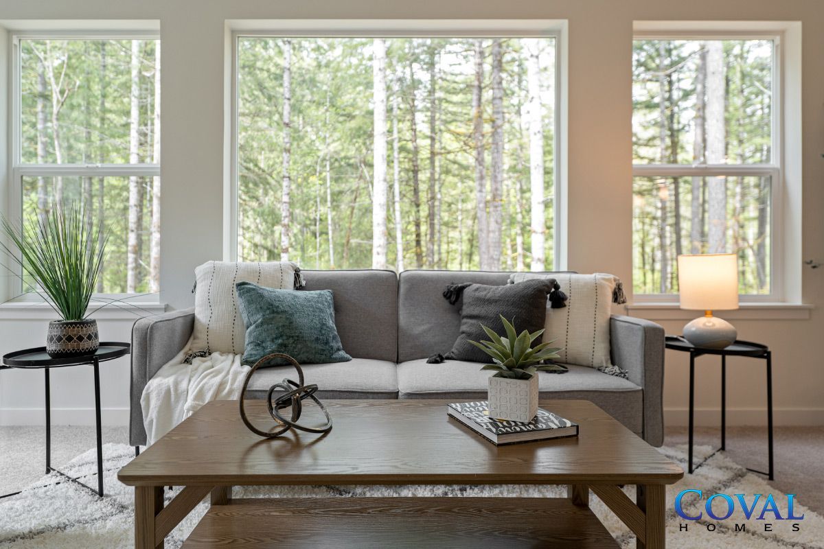 Bright living room with large windows, gray sofa, wooden coffee table, and plants overlooking trees