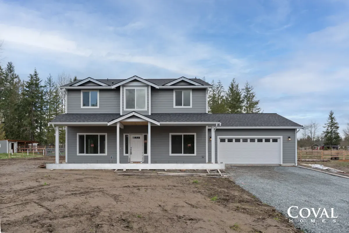 New two-story gray house with white trim, front porch, and attached garage on a dirt lot