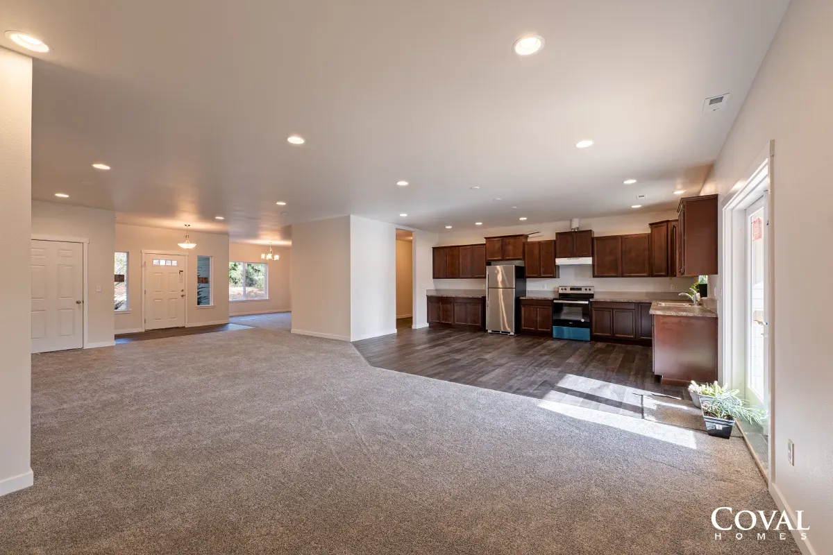 Empty open-concept living room with carpet and recessed lights, leading to a kitchen with dark wood cabinets