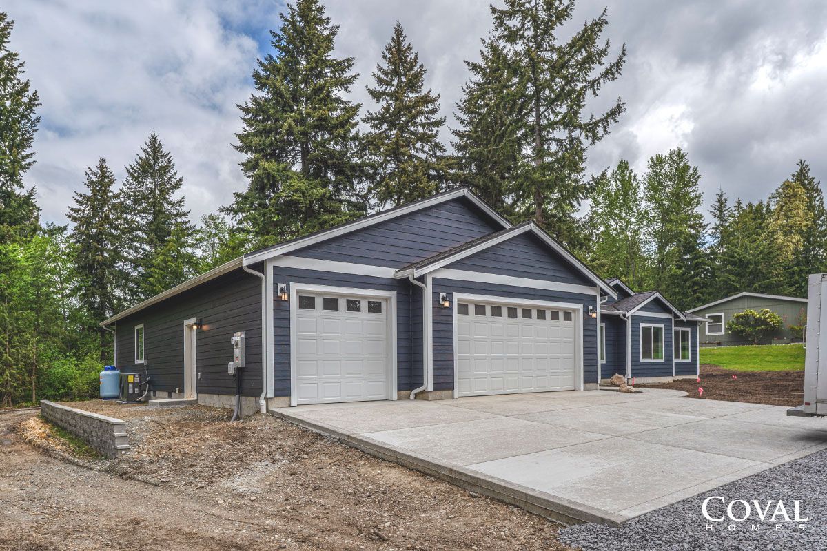 Gray two-car garage with dark blue siding and surrounding trees under a cloudy sky