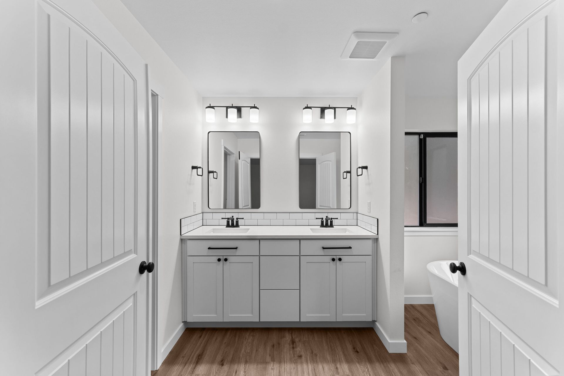 Bright white bathroom with double vanity, mirrors, sconces, and wood floor viewed from the doorway
