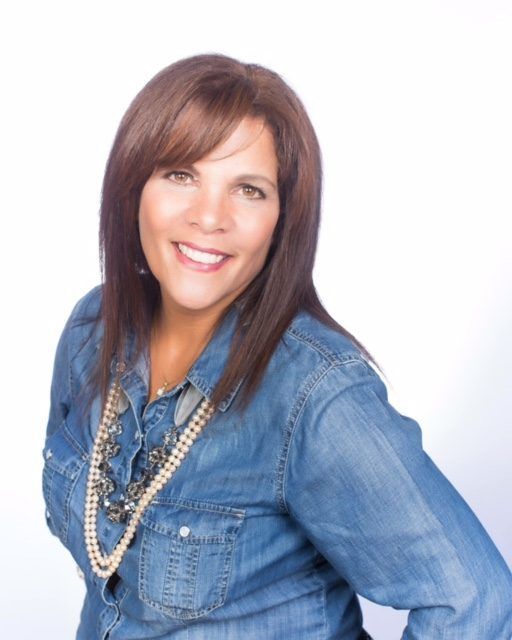 Smiling woman in a denim shirt with layered necklaces against a white background