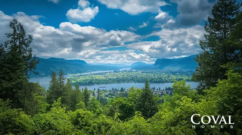 A scenic, high-vantage view of a wide river winding through a lush, forested mountain valley under a bright blue sky filled with puffy white clouds. Green trees frame the foreground, and a town is visible along the riverbanks in the distance. The logo "COVAL HOMES" is displayed in the bottom right corner.