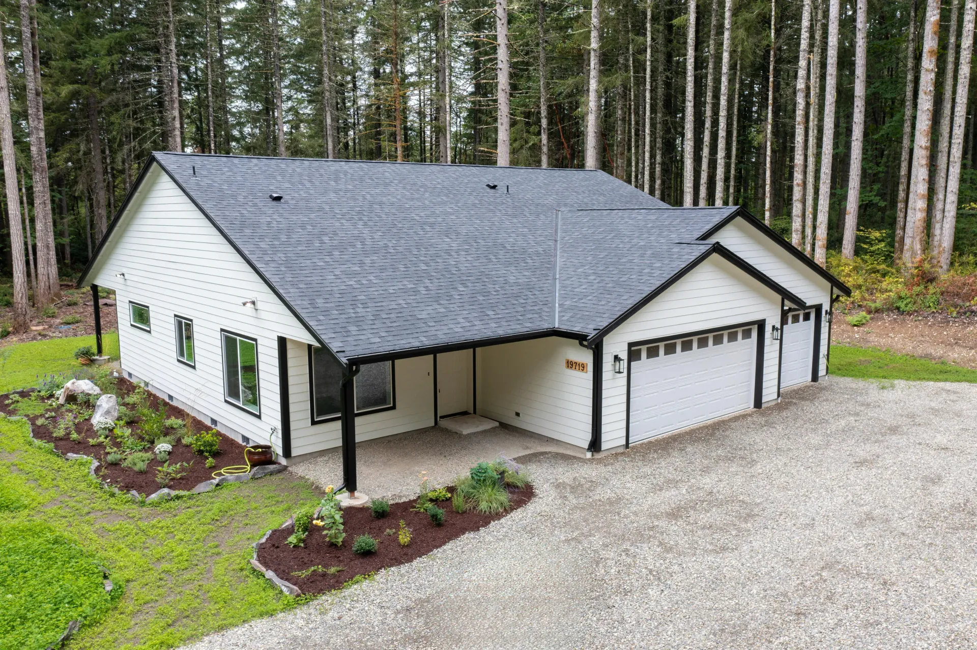 White single-story house with black roof and garage, surrounded by trees and gravel driveway