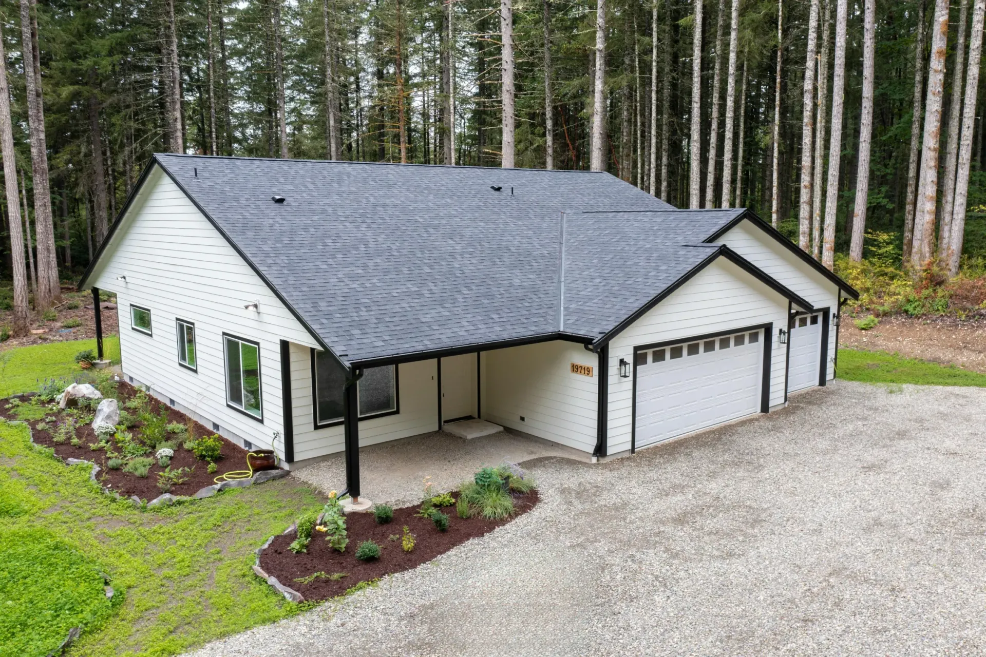 White house with dark gray roof and attached garage, set in a wooded area with gravel driveway.