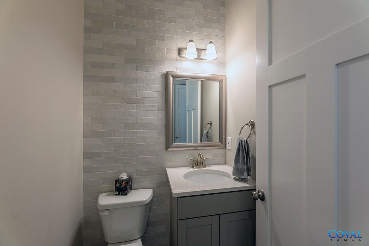Small bathroom with a toilet, vanity sink, mirror, and wall light against gray tile.
