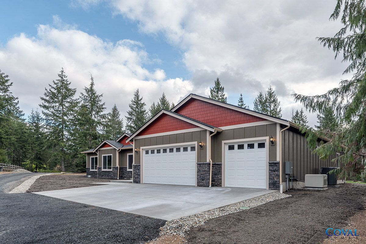 New red-trim house with two white garage doors and a concrete driveway in a wooded setting