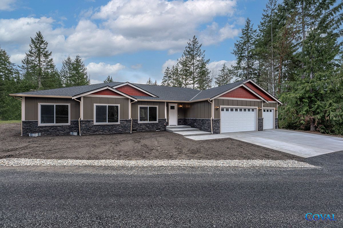 Single-story house with a two-car garage, gravel driveway, and trees under a cloudy sky