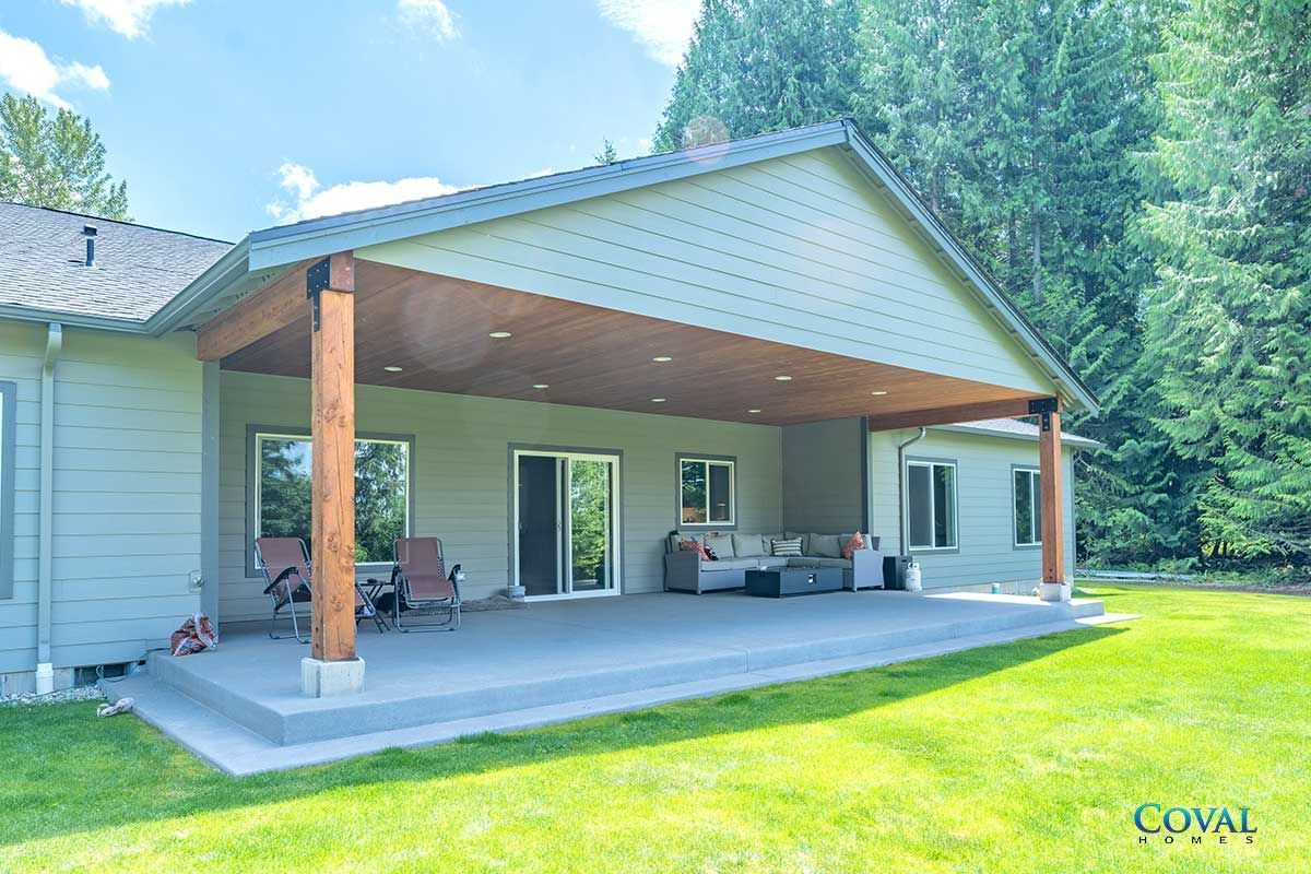 Covered patio with gray seating and wooden posts beside a house, overlooking a sunny lawn and trees