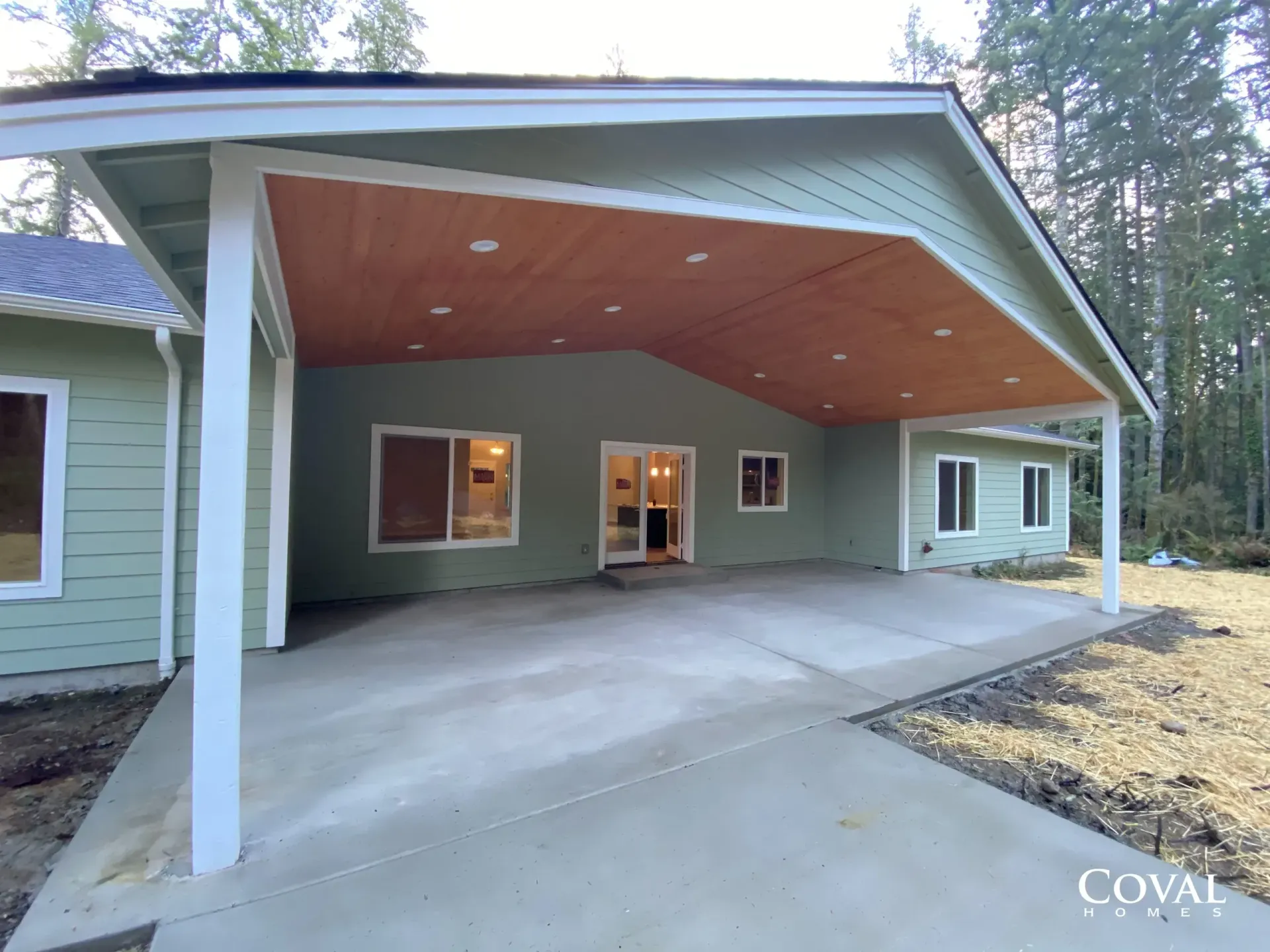 Covered concrete patio with angled wood ceiling attached to a house exterior, with support posts and windows.