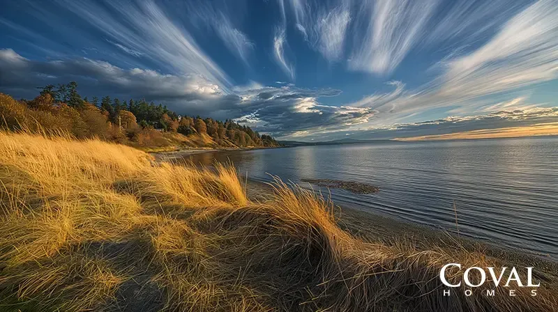 A scenic coastal view featuring tall, golden beach grass growing on sandy dunes in the foreground. Beyond the grass, calm dark water gently laps the shore, which curves to the left and is bordered by a bluff covered in evergreen and autumn-tinged trees. Above, a dramatic deep blue sky is filled with sweeping, feathery white cirrus clouds radiating outward, with a soft golden glow on the distant right horizon indicating sunrise or sunset. The logo "COVAL HOMES" is displayed in the bottom right corner.