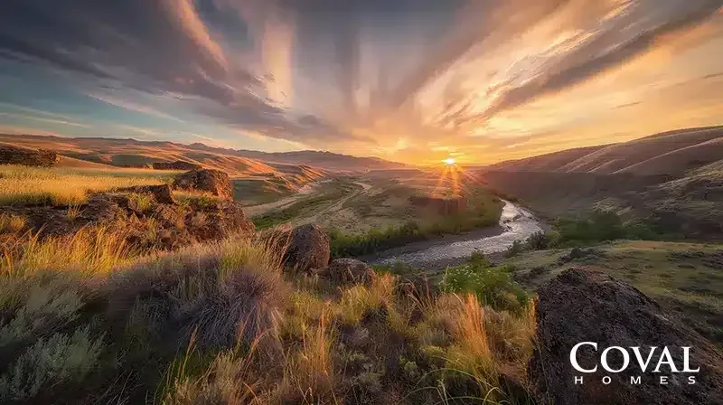 An elevated, scenic view looking down into a deep canyon where a river winds through the valley floor. The foreground features rugged, dark rocks interspersed with tall, golden grasses catching the warm light. In the background, rolling hills stretch toward a brilliant sunrise or sunset, with the sun bursting over the horizon and illuminating dynamic, sweeping clouds in vibrant shades of orange, gold, and soft purple. The logo "COVAL HOMES" is displayed in the bottom right corner.