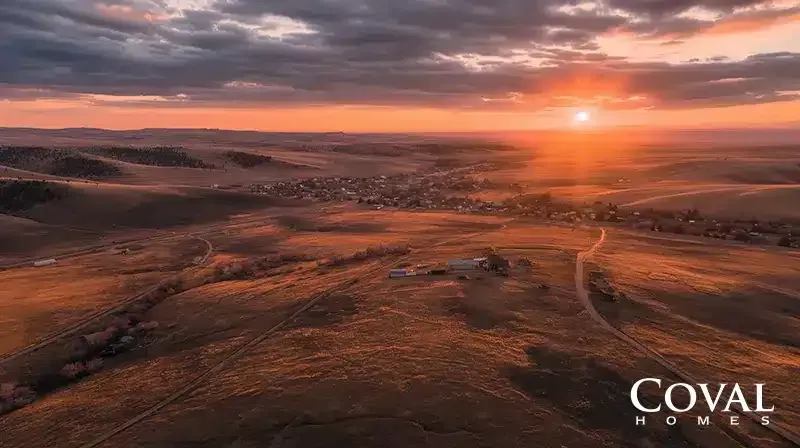 An aerial view of a sweeping, golden-brown landscape of rolling hills bathed in the warm, orange light of a vibrant sunrise or sunset. The bright sun glows low on the right horizon under a dramatic sky filled with dark, textured clouds. In the foreground, winding dirt roads lead toward a solitary farmstead, while a small town is nestled in a valley in the distance. The logo "COVAL HOMES" is displayed in the bottom right corner.