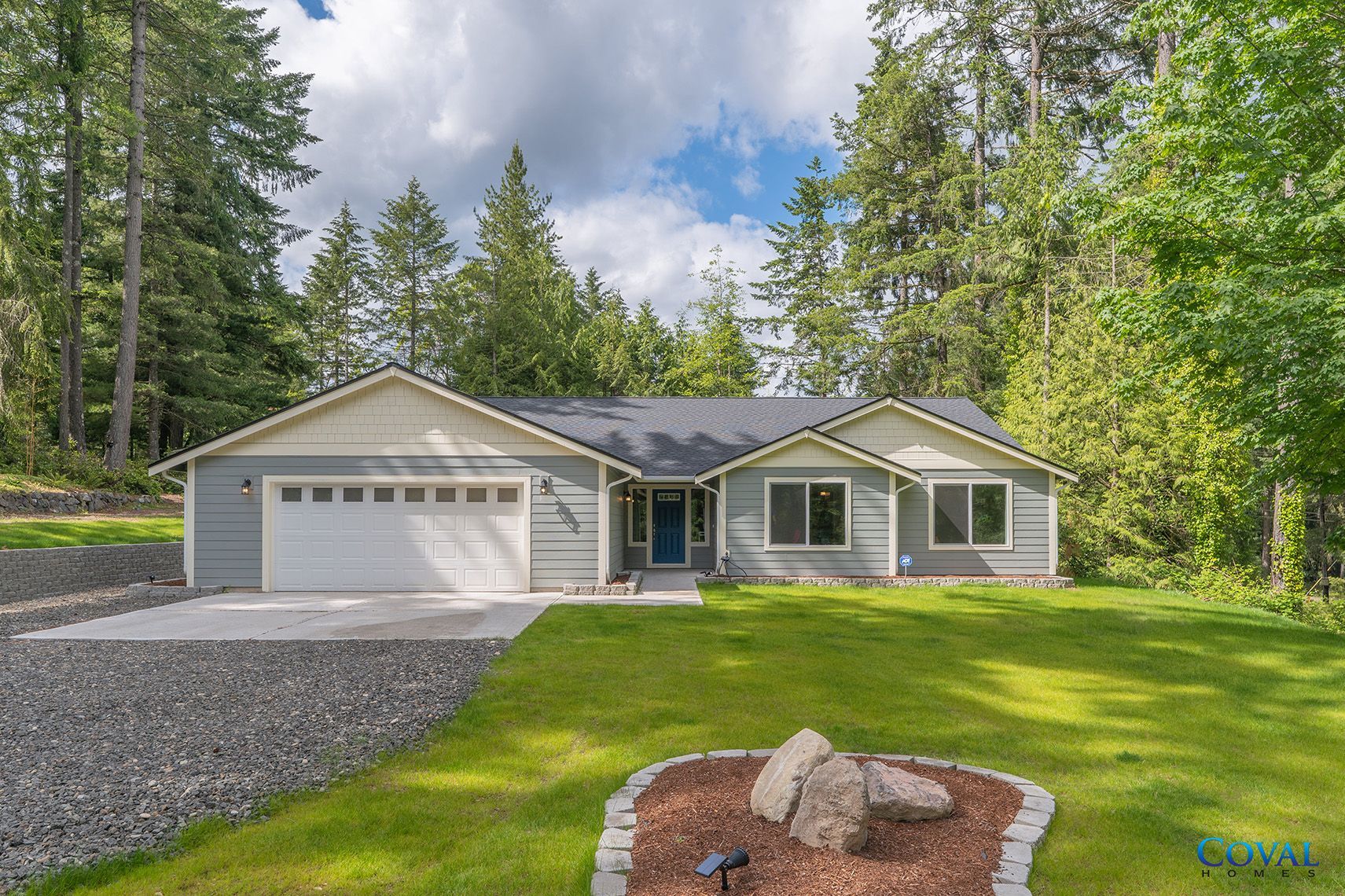 Single-story gray house with attached garage, green lawn, and trees under a partly cloudy sky